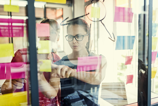Two women stick colourful notes and draw arrows on a glass wall.