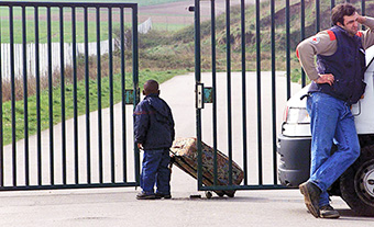 This is a photograph of a young boy holding a suitcase, looking through an open gate.