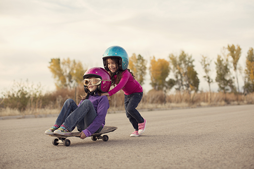 One child sat on a skateboard, another child pushing the skateboard
