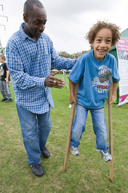 A child walking on stilts with an adult watching close by.