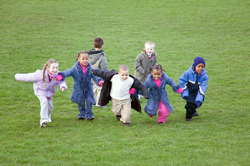 Five children walking hand in hand on grass outside. There are two other children behind them