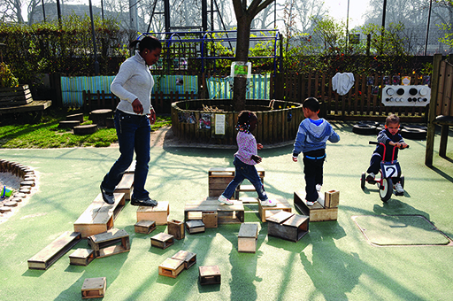 Three children in a playground. One is on a trike, the others are walking on wooden stepping stones with an adult following.