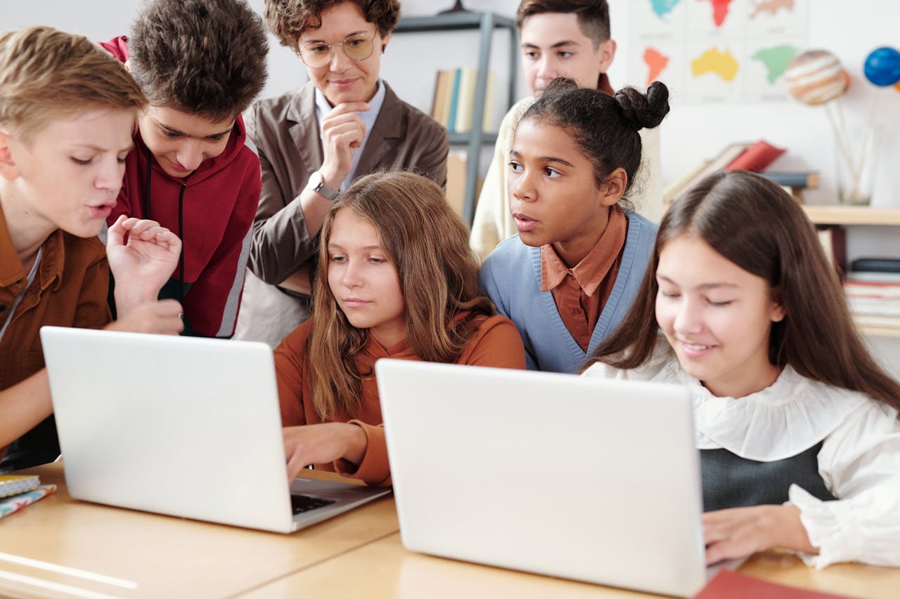School children crowd around two girls sitting at their desks with their laptops.
