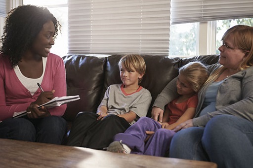 A social care professional, holding a notepad and pen, sits on a sofa beside a mother and her two children.