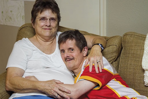 Photograph of a mother seated on a sofa with her adult son. They are both smiling as they sit with their arms around each other.