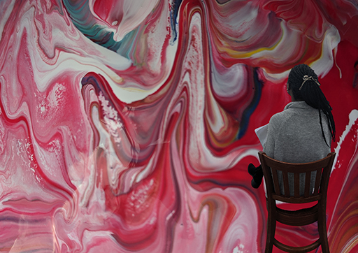 A rear view of the artist Bokani sat on a chair looking at her marbled effect artwork.
