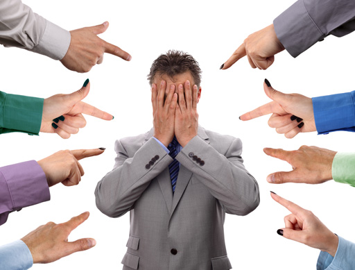 A man in a suit is surrounded by fingers pointing at him. He is covering his face.