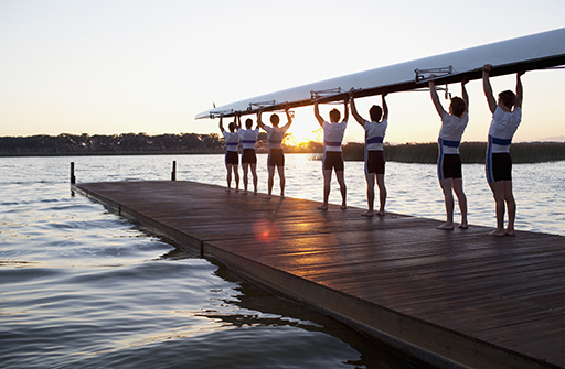 An image showing the backs of seven males holding up a rowing boat above their head.