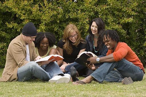 Group of five young adults sat outside looking at two books and smiling.