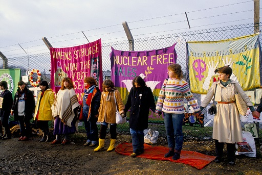 A photograph of a group of women standing hand in hand in front of a number of protest signs.