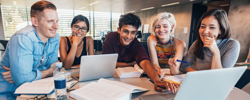Grŵp o fyfyrwyr yn eistedd wrth fwrdd, yn edrych ar liniadur ac yn gwenu