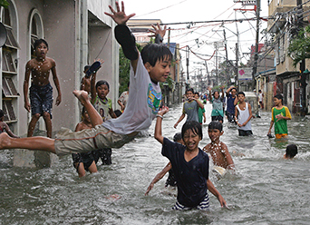 Photo of children playing in a flooded street, one child leaping up in the air.