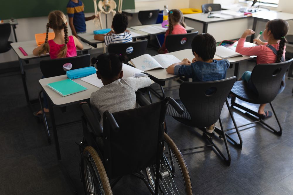 A classroom with 5 students pictured at desks. One student is a wheelchair user.