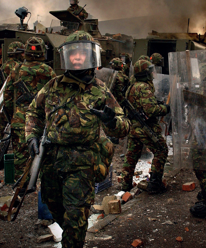 Photo of a group of armed people in camo uniform and riot gear. Some armoured vehicles in the background. The ground is littered with bricks and debris. One of the armed officers turns to the camera, saying something and gesturing.