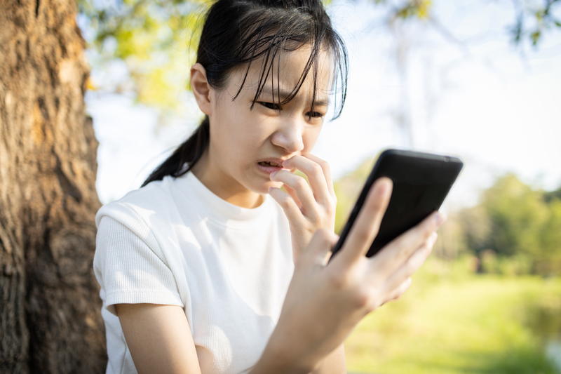 Worried teenage girl - a victim of cyber bullying - sitting alone outdoors while looking at mobile phone