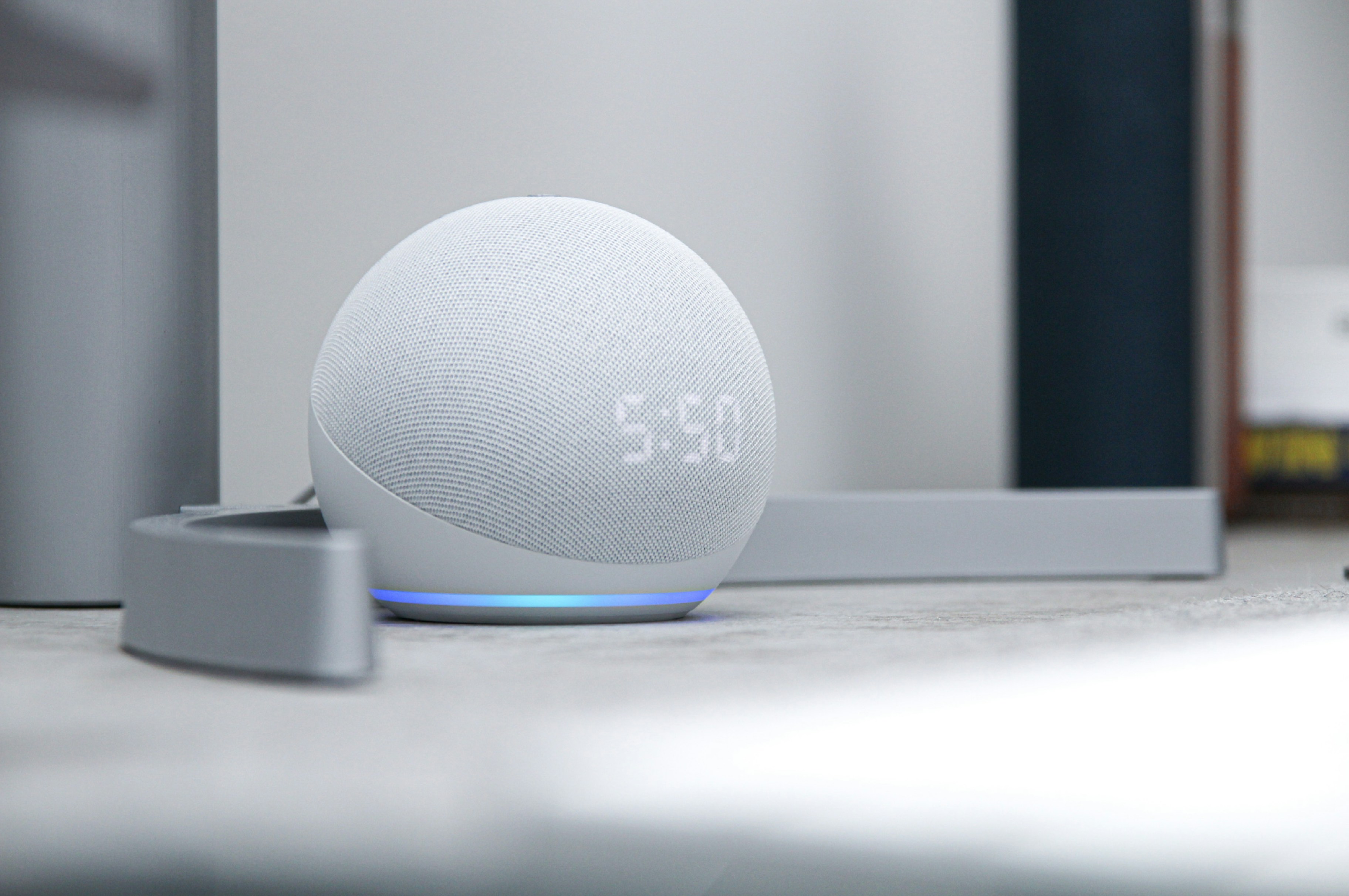 A grey round SMART home device that is displaying the time, sat on a white table.