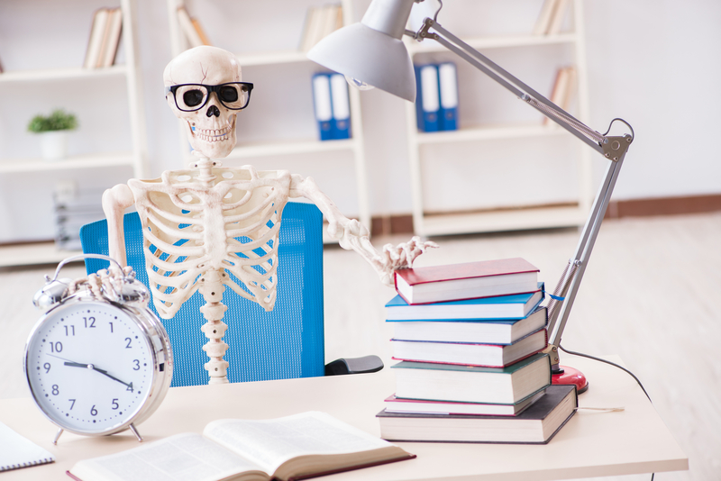 Halloween student skeleton preparing for exams at a desk with books, a notepad and wearing glasses.
