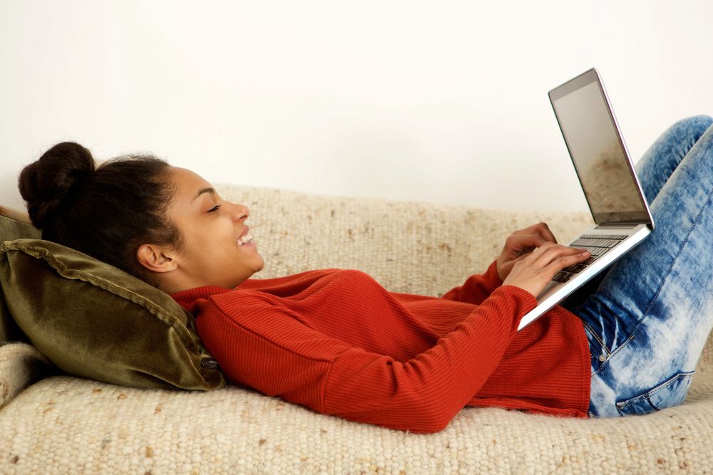 Female student lying on a sofa and typing on laptop