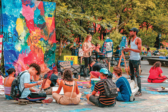 A colour image of a group of young people sitting and standing outdoors. In the background is a large colourful banner painted with the words ‘Black Lives Matter’ on it.