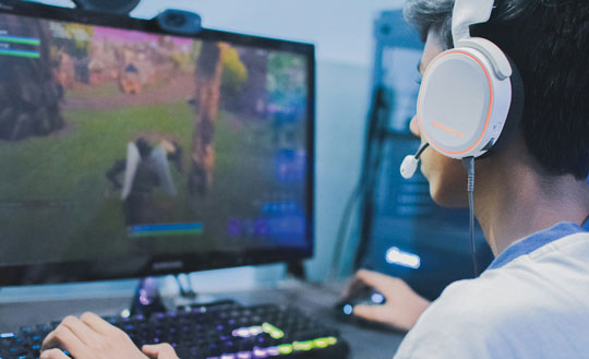 A colour image of a young person wearing headphones, with their hands on a computer keyboard and a game showing on the computer monitor.