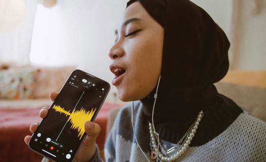 A colour image of a young person wearing a black hijab, grey jumper and several necklaces. She has her mouth open and is singing into a mobile phone which is digitally recording the sound.