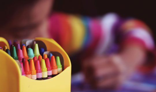 A colour image focused on a yellow pot containing a range of coloured crayons. A child can be seen drawing in the blurred background.