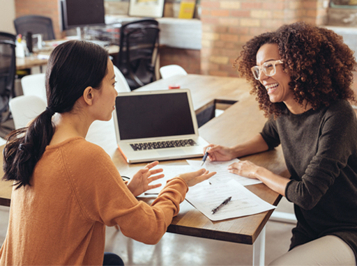 A photograph of two people in an office environment talking to each other.