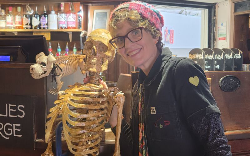Jennie E. Owen smiles at the camera while posing beside a decorative human skeleton in a pub. Another skeleton, resembling a dog, is visible behind them, with bottles lining the bar.