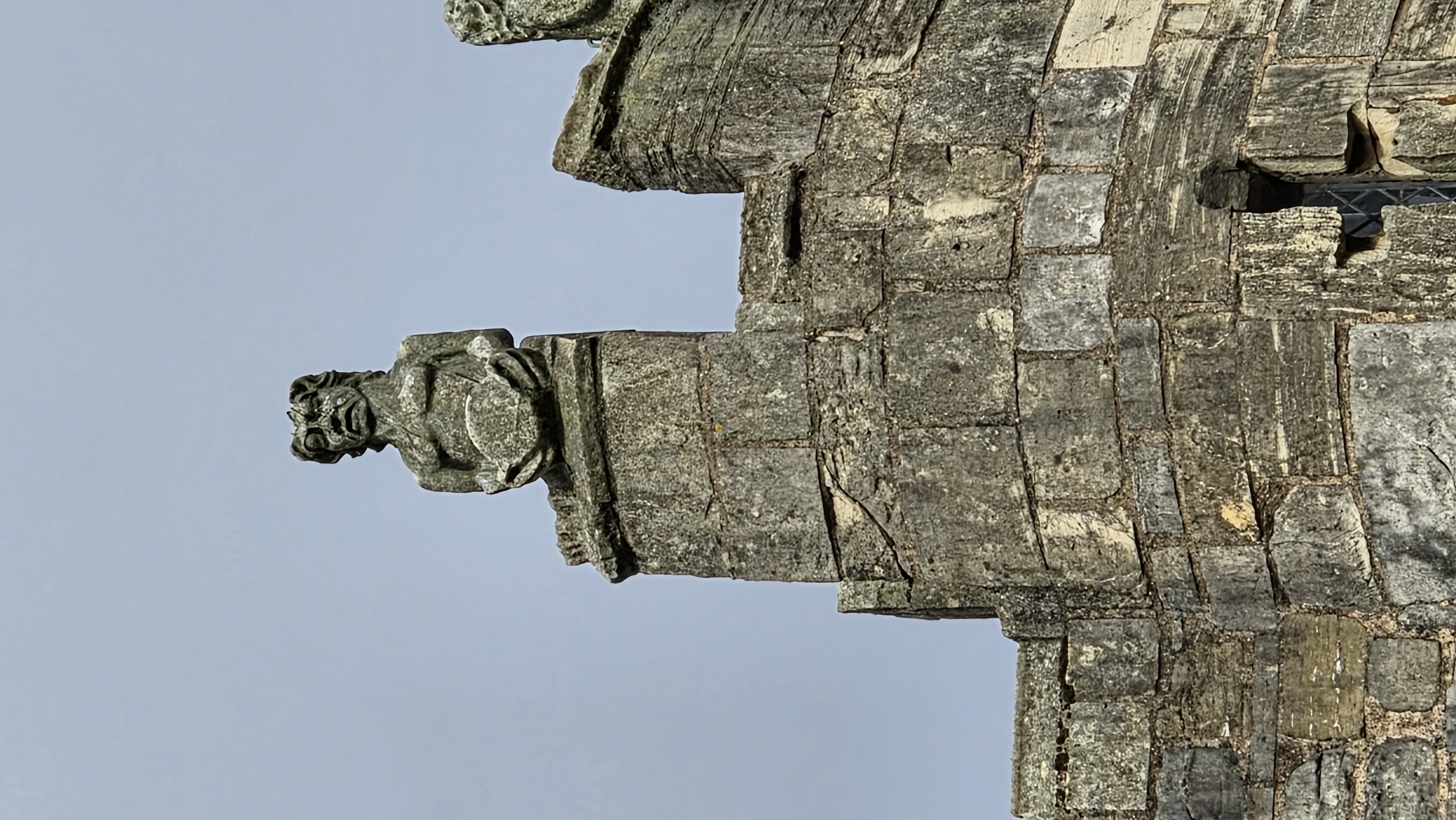 A weathered stone carving of a grotesque holding a circular object sits atop the parapet of an old stone building against a pale blue sky.