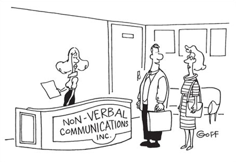 This is a cartoon image. It shows an office reception area. A receptionist stands behind a desk. A sign on the front of the desk reads ‘Non-Verbal Communications Incorporated’. A man and woman stand in front of the desk. The man is speaking to the woman. The caption of the cartoon reads. ‘See? That means, “What do you clowns want?”’