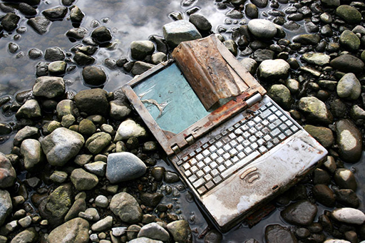 This image shows a rusty laptop on top of some rocks near water.