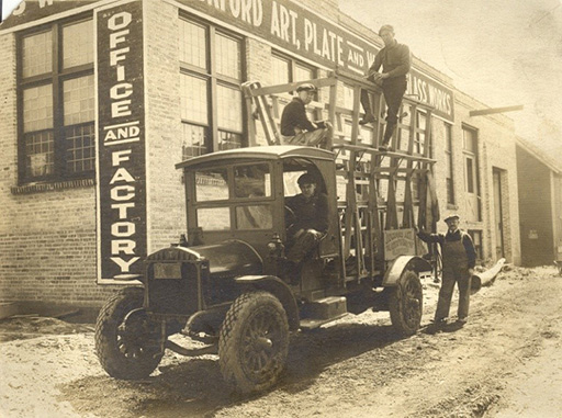 An image of an old black and white photograph of a truck outside a small factory.