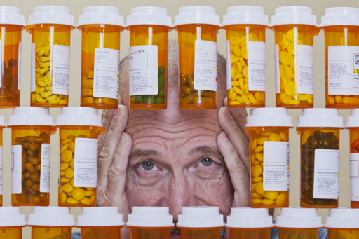 An older man clutches his head in dismay at the bottles of prescription medication surrounding him.
