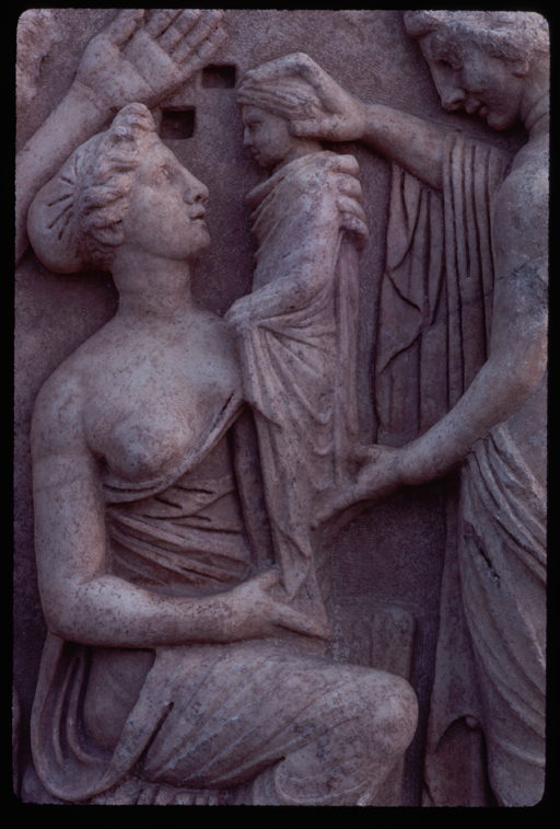 Carved relief of a mother holding a child who is wrapped in swaddling clothes, while another woman supports the child’s head and back.