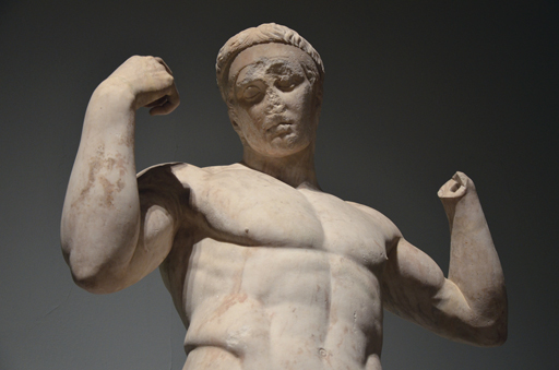 Ancient classical statue of an athlete with his head looking down, and his arms bent and raised at shoulder height.