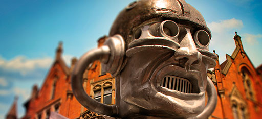 A metal robot face seeming to grimace menancingly in front of a Victorian school building