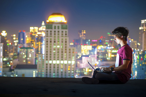 The image is of a young man sitting on a ledge/roof, using a laptop.