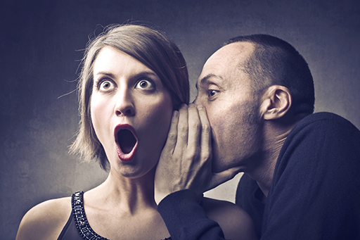 Image of a man whispering into a woman’s ear. The woman has a shocked expression on her face.