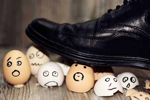 A shiny, black business shoe is crushing several egg shells with worried faces drawn on them.