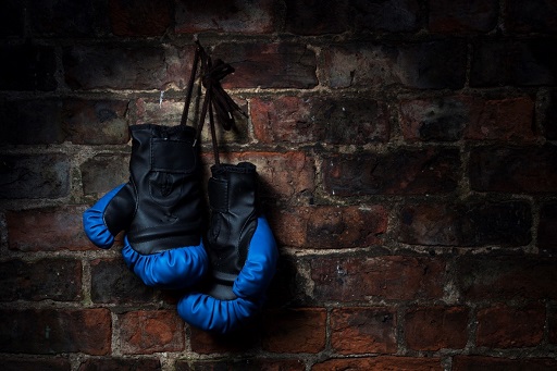 A pair of blue and black boxing gloves hangs against a dark brick wall.