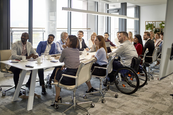 A group of people seated around a conference table in a modern office meeting room, listening to a presentation. Large windows provide natural light.
