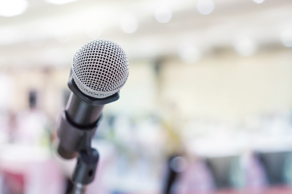 Close-up of a microphone on a stand with a blurred indoor event or presentation space in the background.