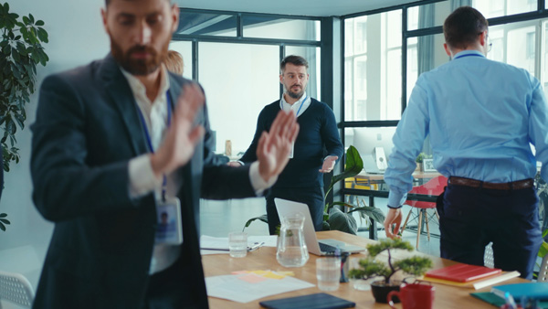 Several people in a modern office meeting room stand around a conference table, with one person in the foreground holding up both hands appearing to start walking out of the room while another stands near the table with open hands. A laptop, notebooks, and glassware are visible on the table.