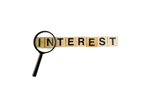 Magnifying glass highlighting the word ‘INTEREST’ spelled with wooden letter tiles on a light background.