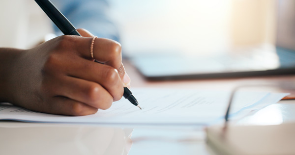 Close-up of a hand holding a pen and signing or writing on a document on a desk, with a blurred laptop in the background.