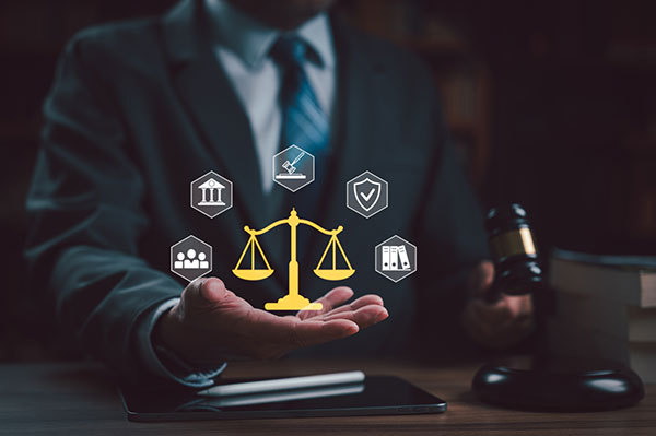 Person in a business suit holding out a hand with a digital scales-of-justice icon and legal symbols, with a judge’s gavel on a desk in the background.