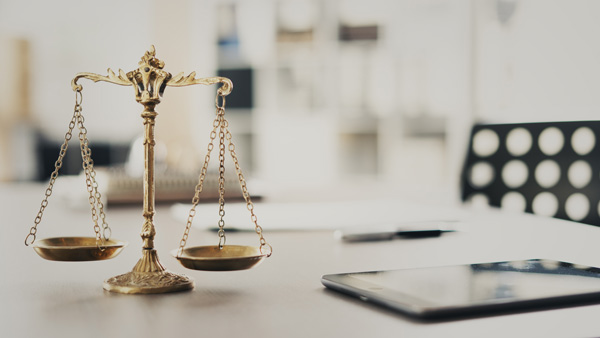 Gold scales of justice on a desk in a modern office, with a tablet and blurred background furnishings.