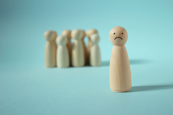 Wooden peg figure with a sad face standing apart from a blurred group of similar figures against a light blue background.