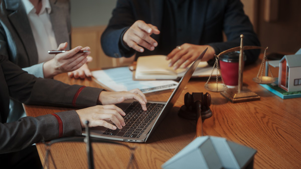 People working around a laptop at a meeting table with legal and real-estate symbols, including a gavel, scales of justice, documents and model houses.