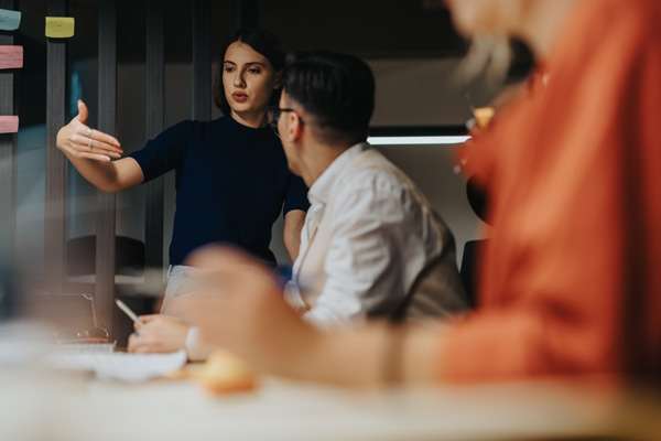 Person presenting ideas to colleagues in a modern office, gesturing toward a wall with sticky notes while others listen around a table.